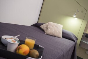 Una cama bien hecha con un edredón gris y una almohada blanca, una bandeja con desayuno que incluye un tazón de cereales, un vaso de jugo de naranja, un melocotón y un kiwi.