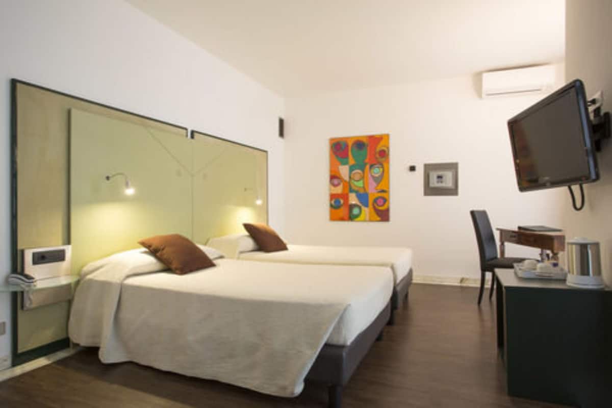Ein Hotelzimmer mit zwei Betten, einem Schreibtisch, einem Sessel, einem Fernseher und einem farbenfrohen abstrakten Gemälde an der Wand.