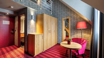 Une chambre d’hôtel avec un tapis rouge, une armoire en bois, une table ronde ornée d’une plante en pot, un fauteuil rouge et une lampe rouge.