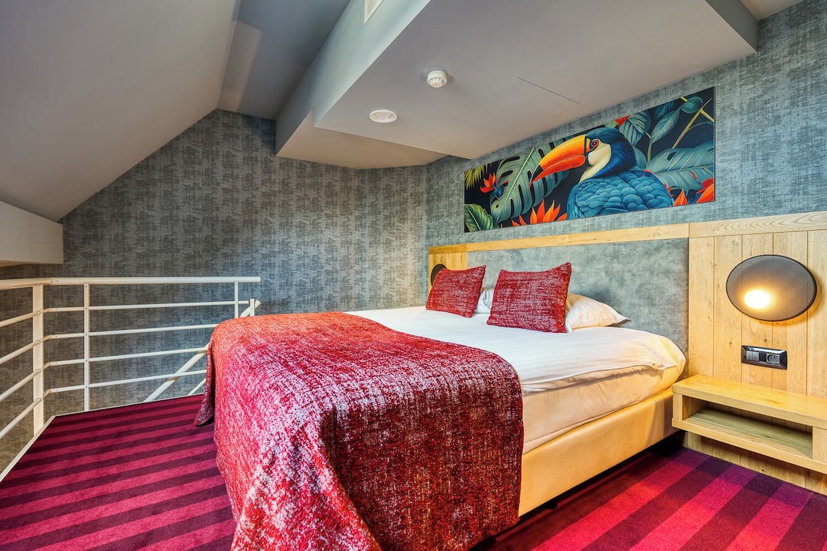 Een moderne slaapkamer met een groot bed, rode beddengoed, een houten hoofdeinde, een wandlamp en een tropische muurschildering.