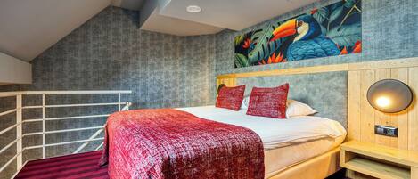 Une chambre moderne avec un grand lit, une literie rouge, une tête de lit en bois, une lampe fixée au mur et une fresque murale aux motifs tropicaux.