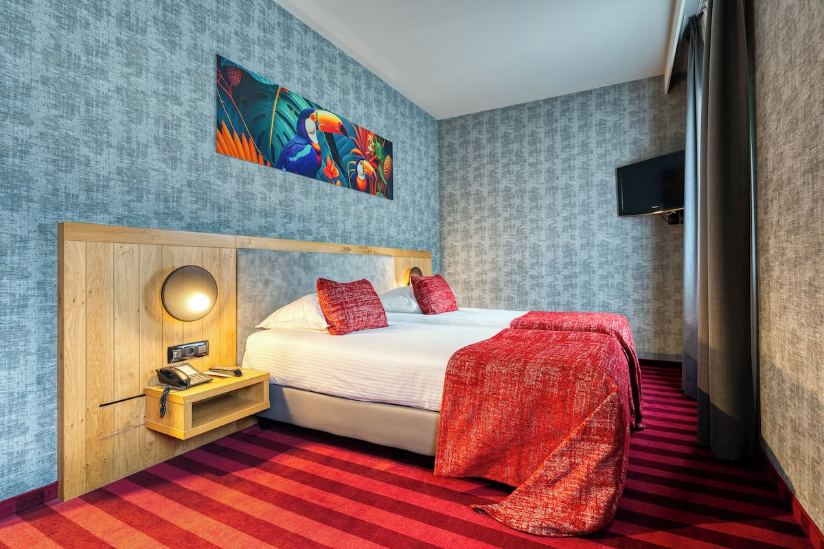 Een hotelkamer met een groot bed, een houten nachtkastje met een lamp, een rood-wit gestreept tapijt en een kleurrijk abstract schilderij aan de muur.
