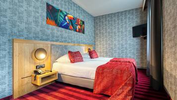 A hotel room with a large bed, a wooden bedside table with a lamp, a red and white striped carpet, and a colorful abstract painting on the wall.