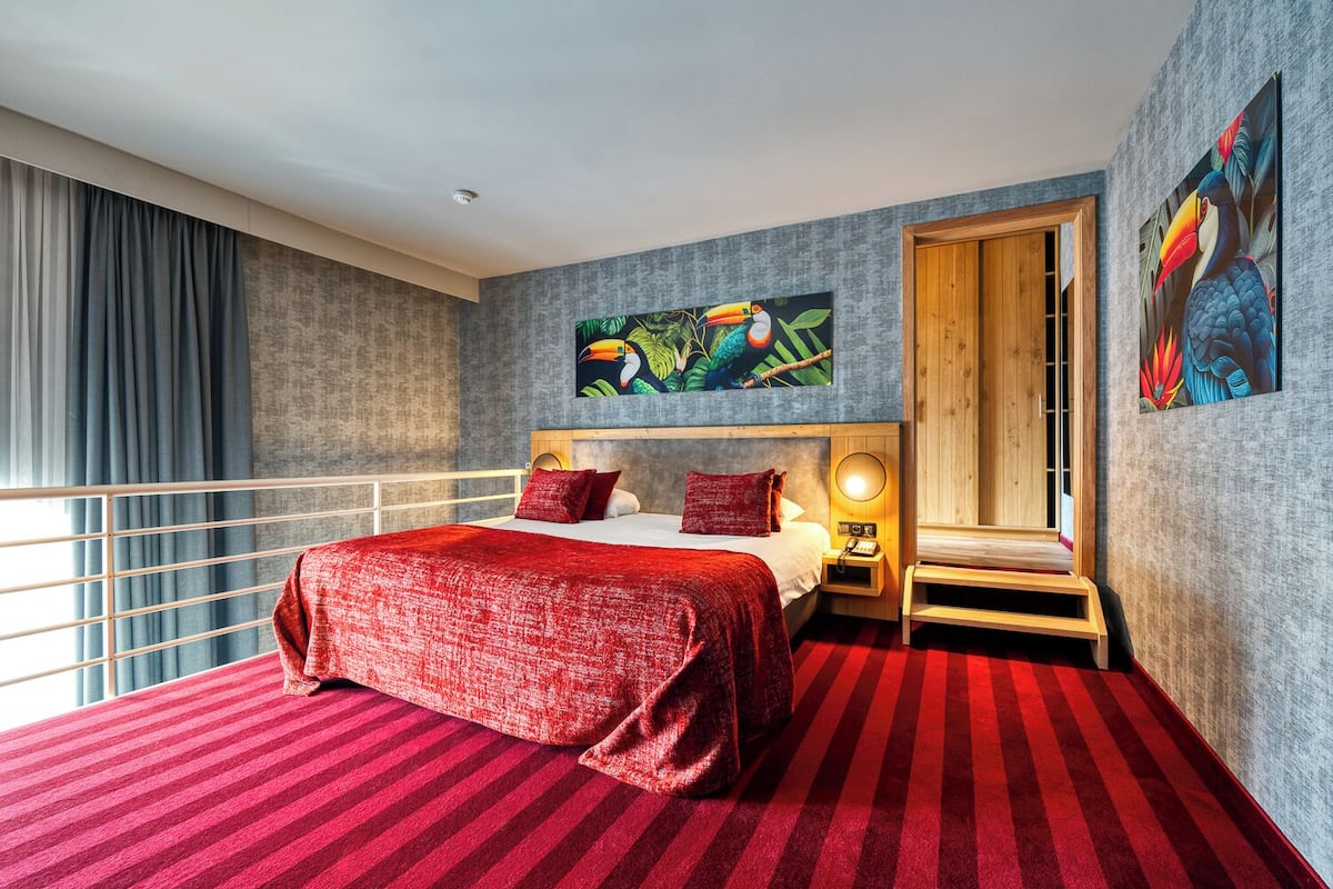 Een hotelkamer met een groot bed, een houten nachtkastje, een rood-gestreept tapijt en een raam met gordijnen.