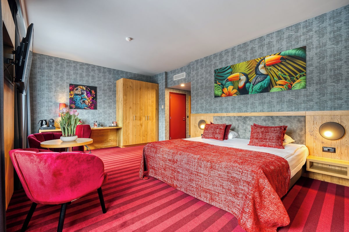 Hotelkamer met een groot bed, een rood gestreept tapijt, een rode stoel, een houten garderobe, een klein tafeltje met een plant en een schilderij van toekans aan de muur.
