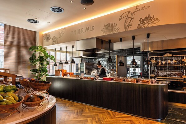 A modern kitchen with a chef preparing food, a bar area, and a counter with fruit bowls. - Hotel Verviers Van der Valk (Verviers)
