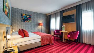 A hotel room with a bed, a desk, two chairs, a lamp, and artwork on the walls.