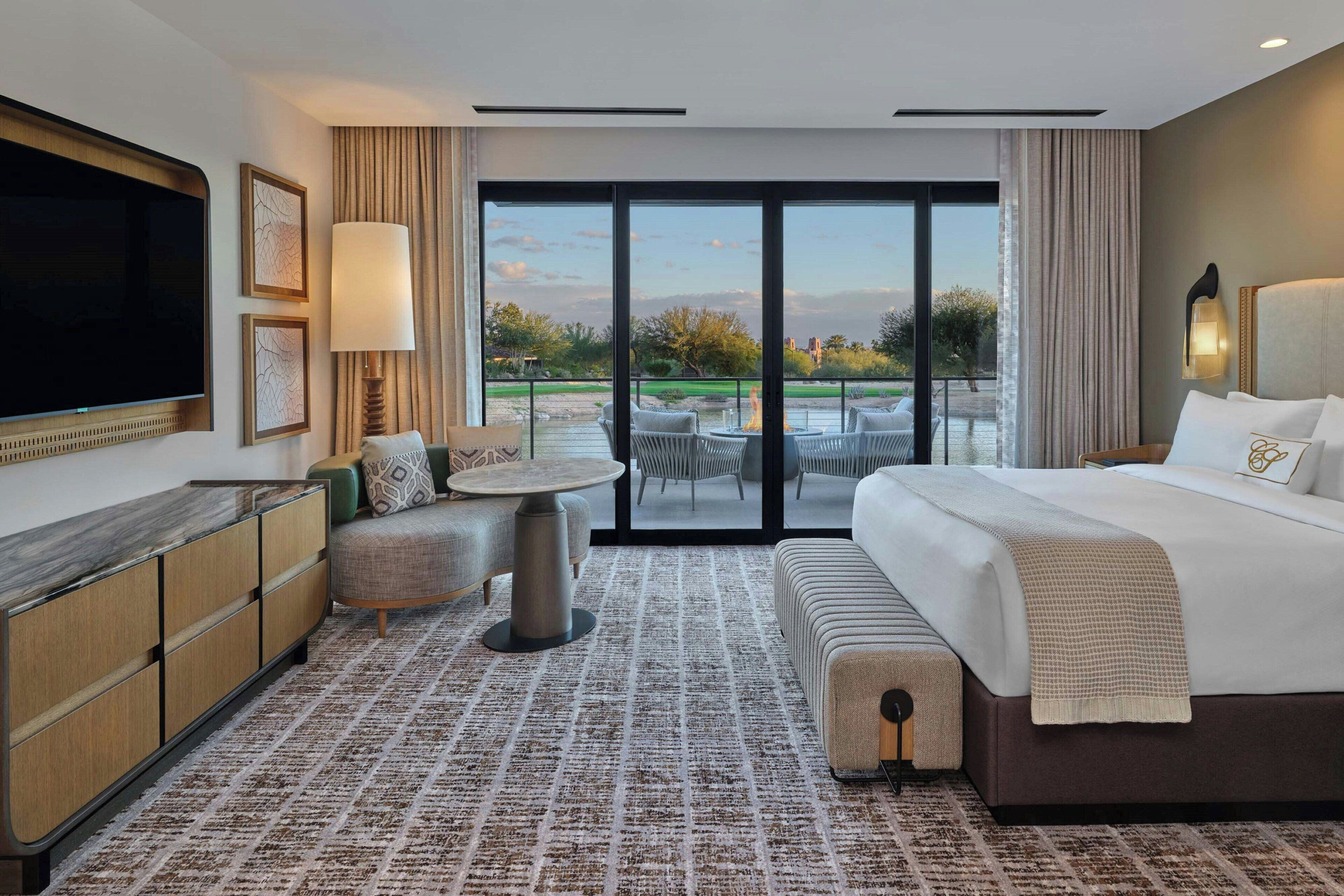 Une chambre d’hôtel avec un grand lit, une télévision, un bureau et une vue sur une terrasse extérieure aménagée avec des tables et des chaises.