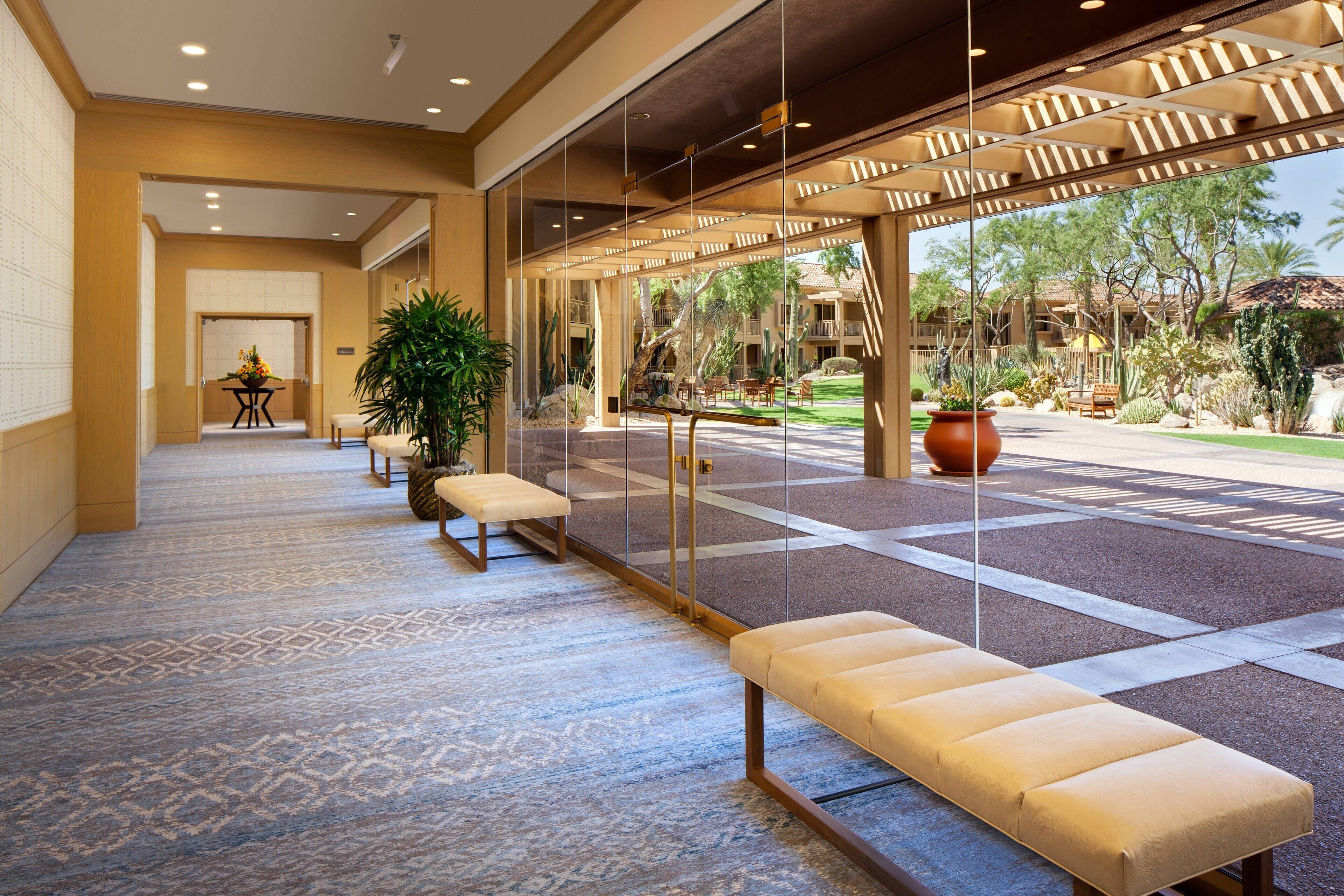 A spacious lobby with a patterned carpet, a seating area with benches, and a view of the outdoor garden through large glass doors.