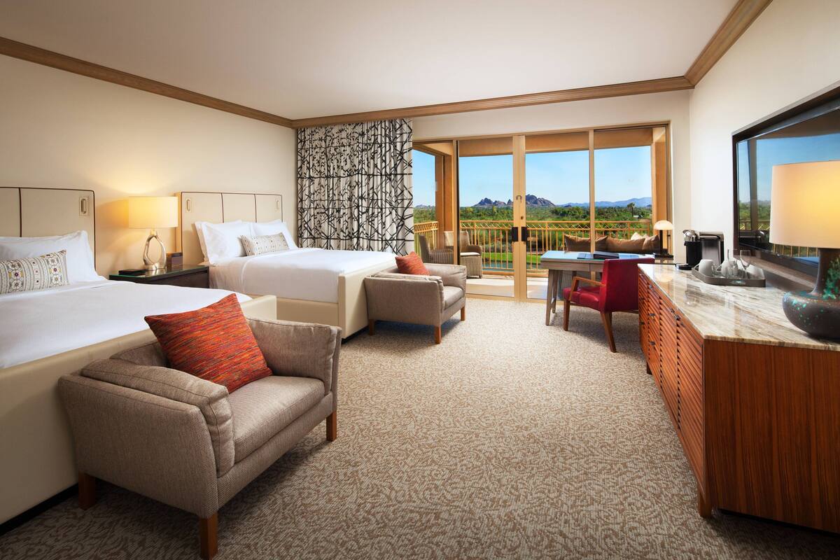 A hotel room with two beds, a sofa, a desk, and a large window with a view of mountains.