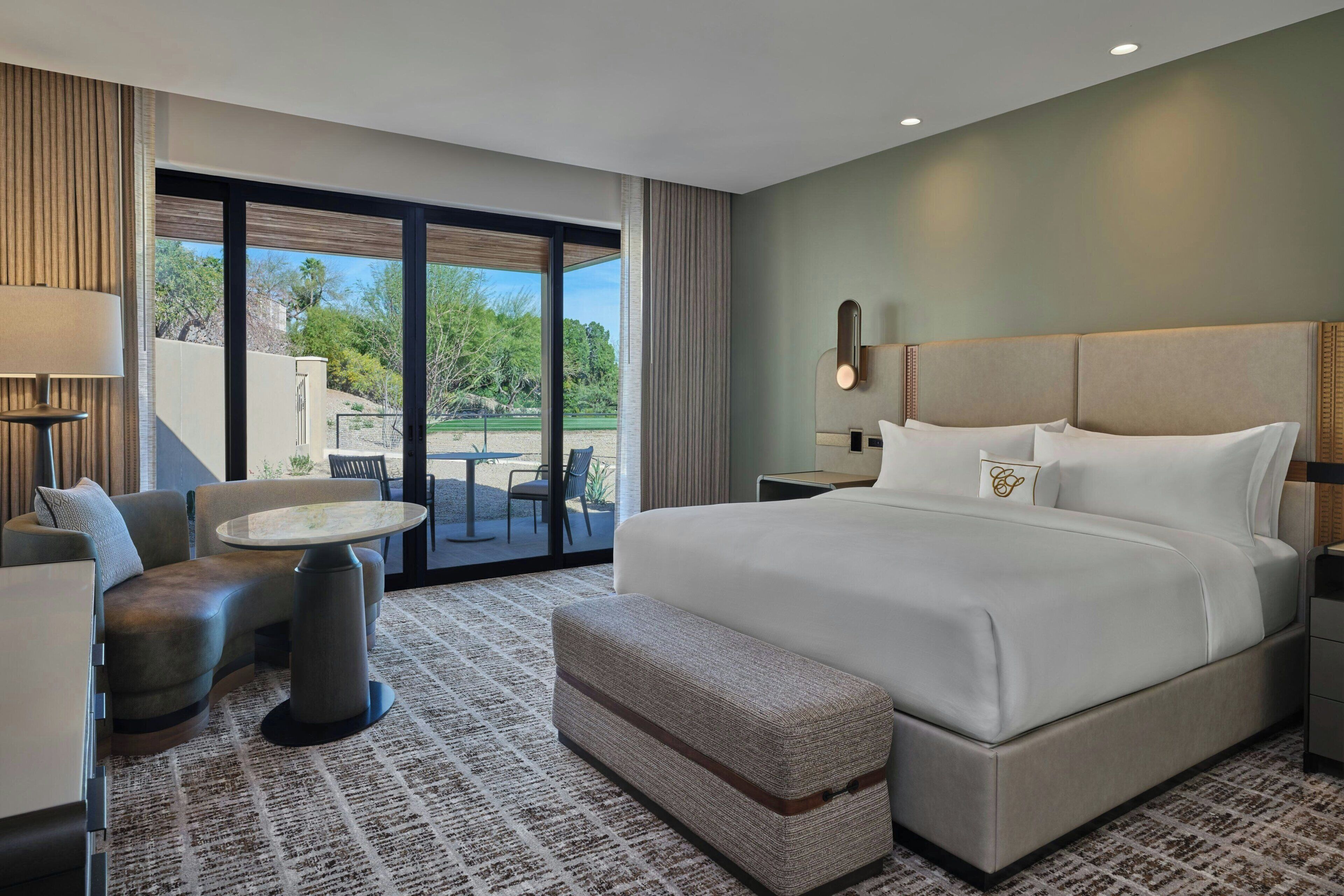 A hotel room with a large bed, a seating area with a table, and a view through a glass door to an outdoor area.