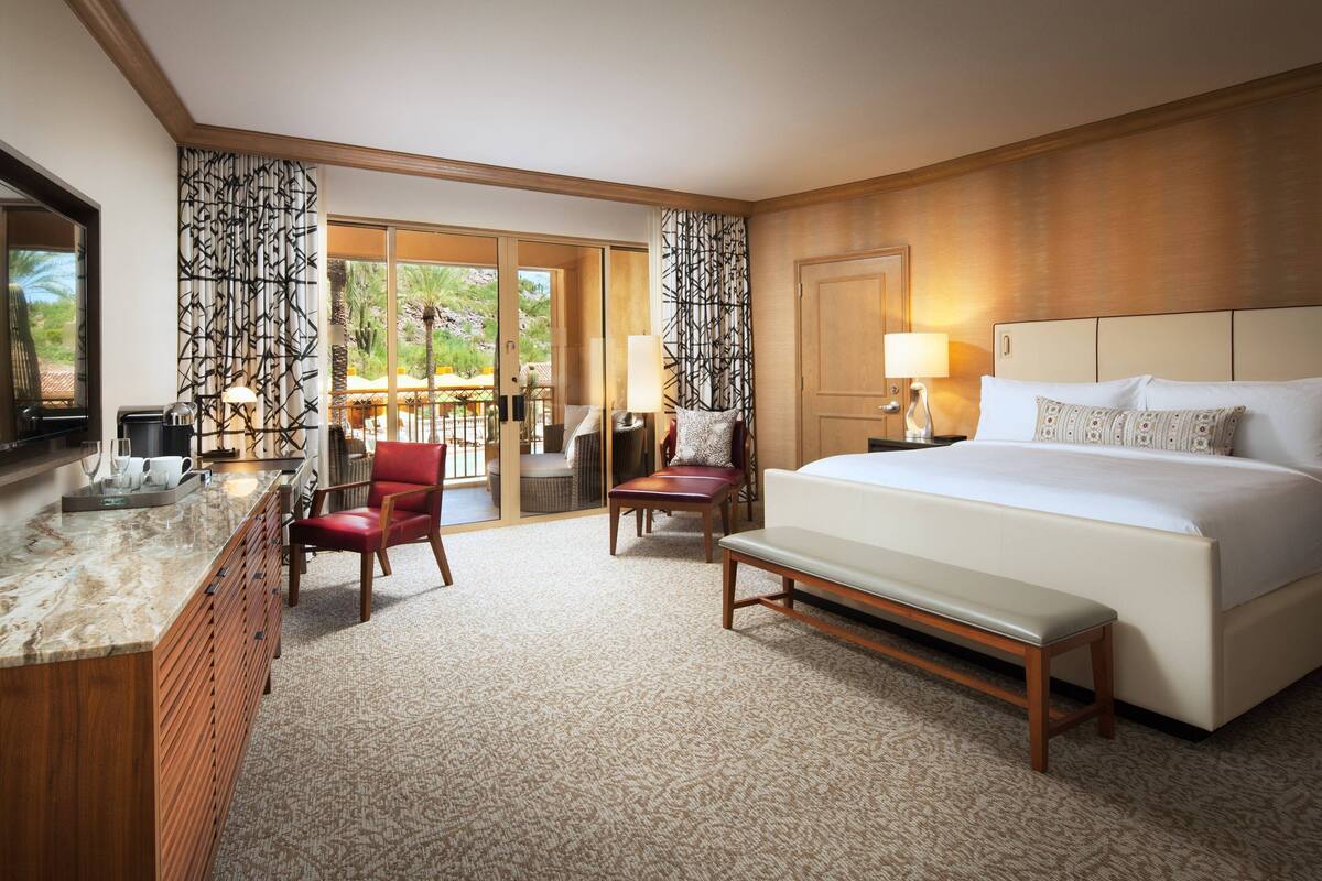 A hotel room with a large bed, a desk with a chair, a bench, and a view of the outdoors.