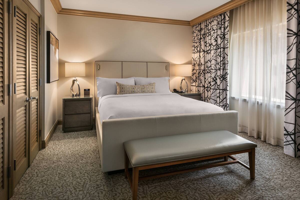 A hotel room with a large bed, a bench, a nightstand, a lamp, and a patterned curtain.