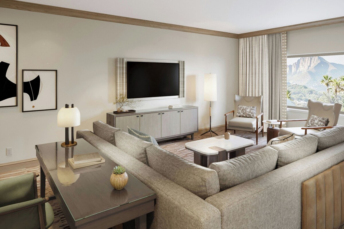 A modern living room with a sofa, coffee table, TV, and a view of a mountain landscape.