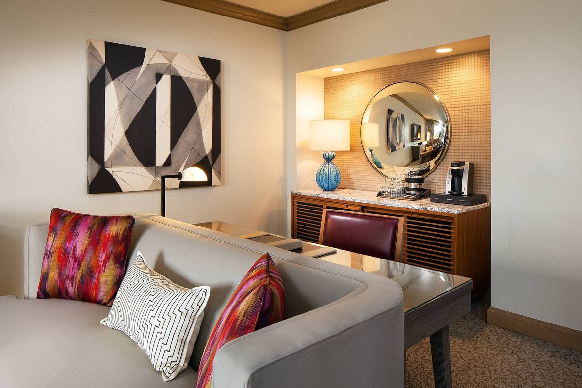 A hotel room with a sofa, a coffee machine, a mirror, and a geometric art piece on the wall.