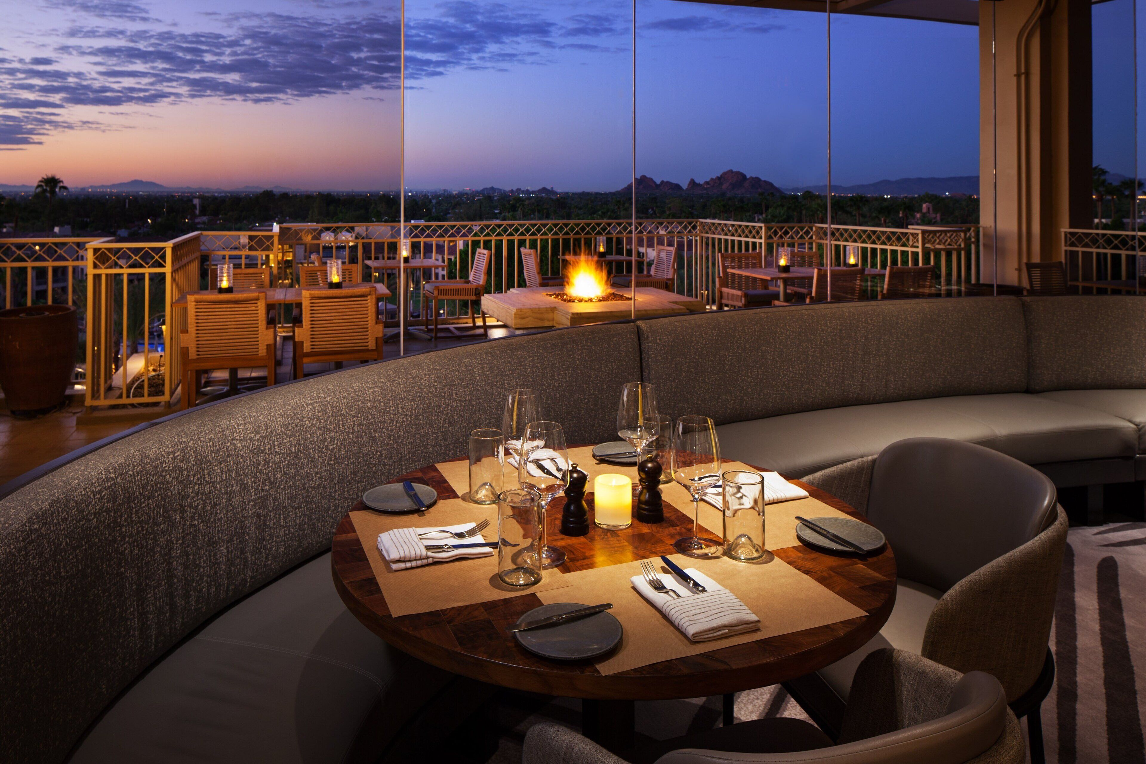 A dining area with a round table set for four, surrounded by cushioned seating. A fire feature is centrally placed on the table, and the setting overlooks a scenic view of a cityscape and mountains.