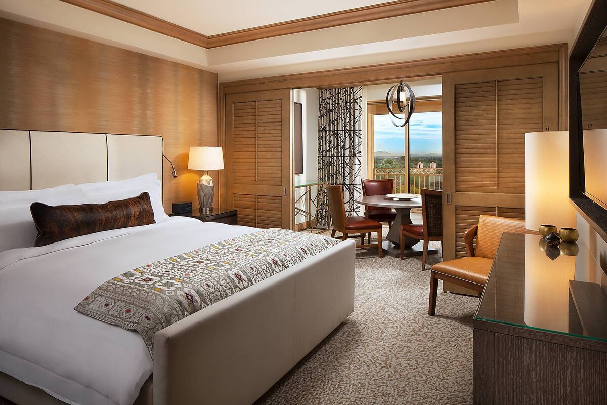 A hotel room with a large bed, a desk with a chair, a TV, and a balcony with a view.