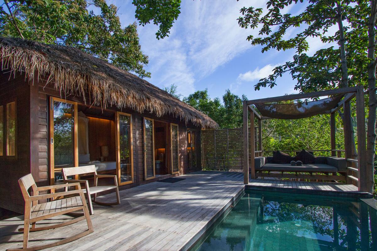 A wooden house with a thatched roof, a pool, and a wooden deck with seating.