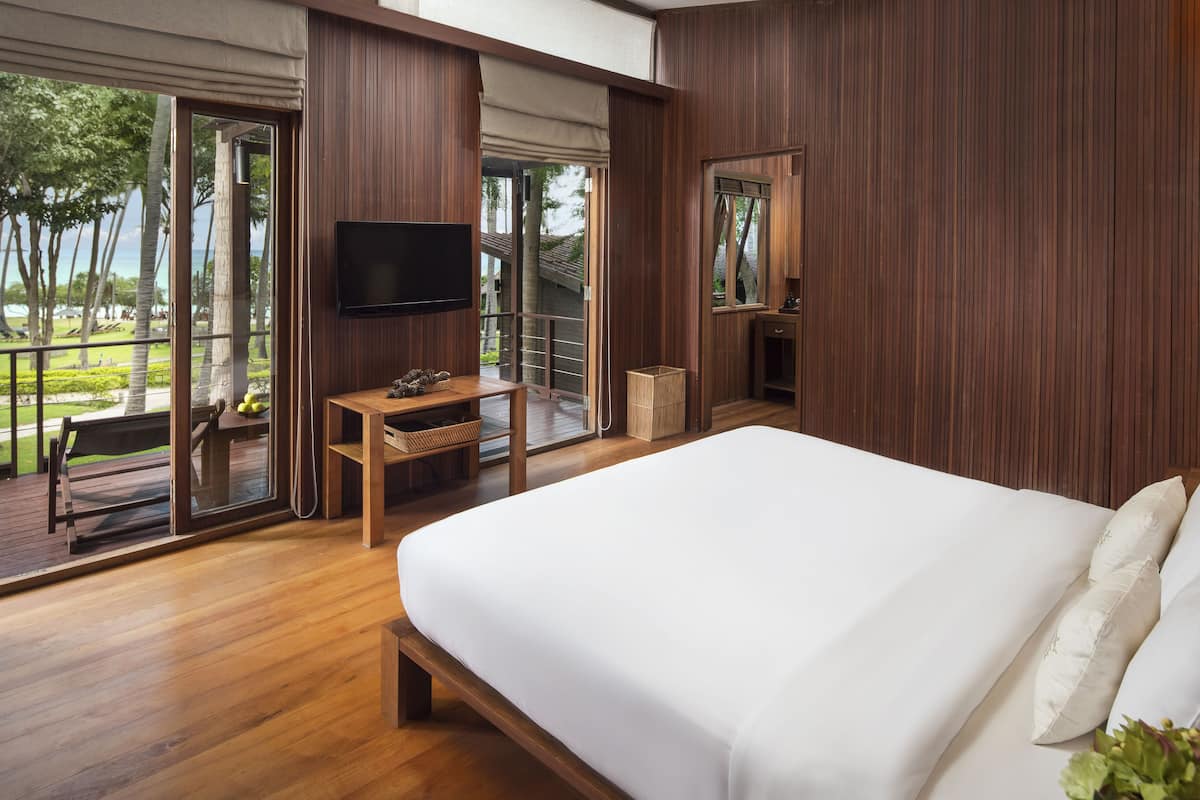 A bedroom with a large bed, wooden flooring, a TV, and a balcony with a view of greenery.