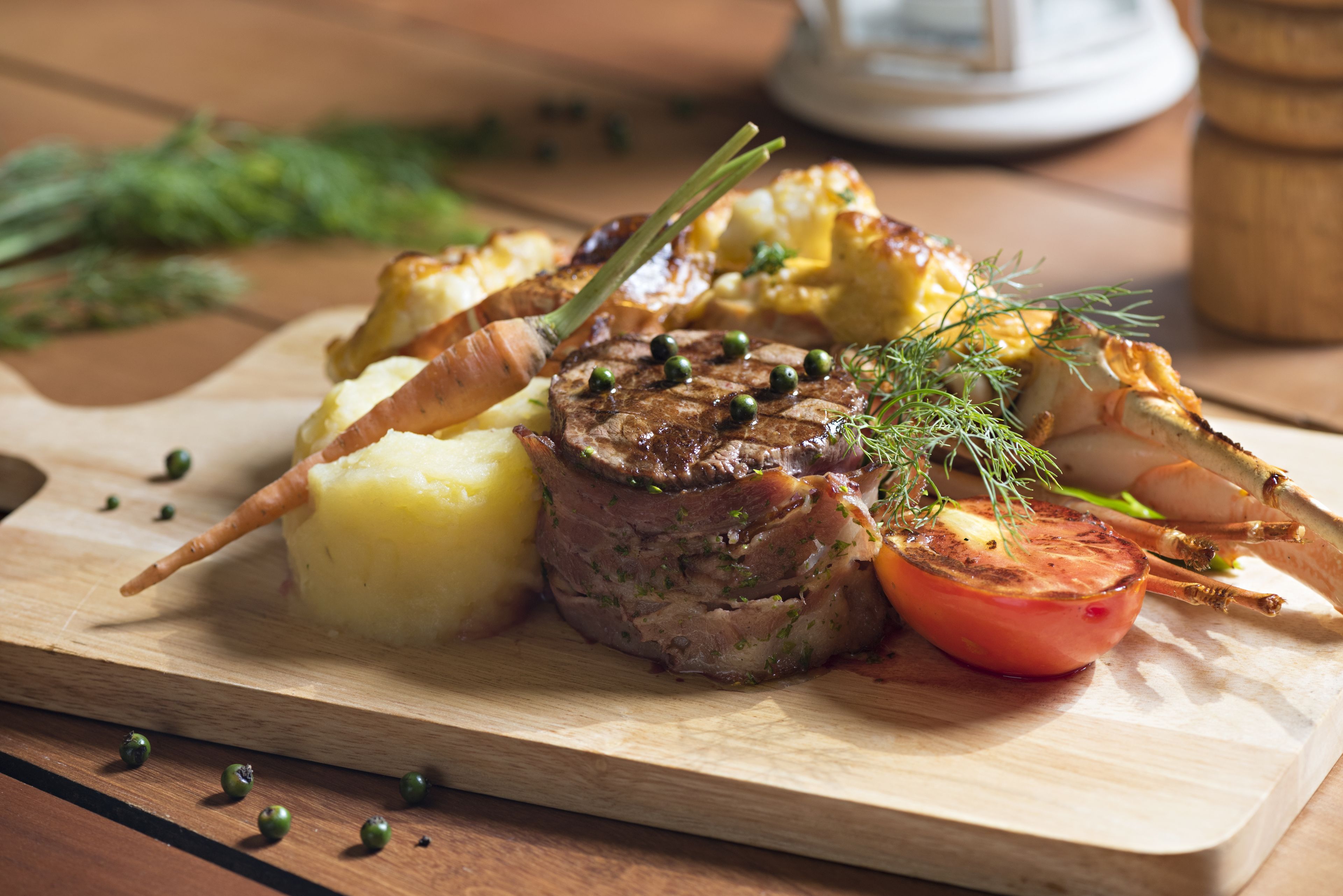 A wooden cutting board with a roasted beef steak, potatoes, and a tomato on a wooden table.