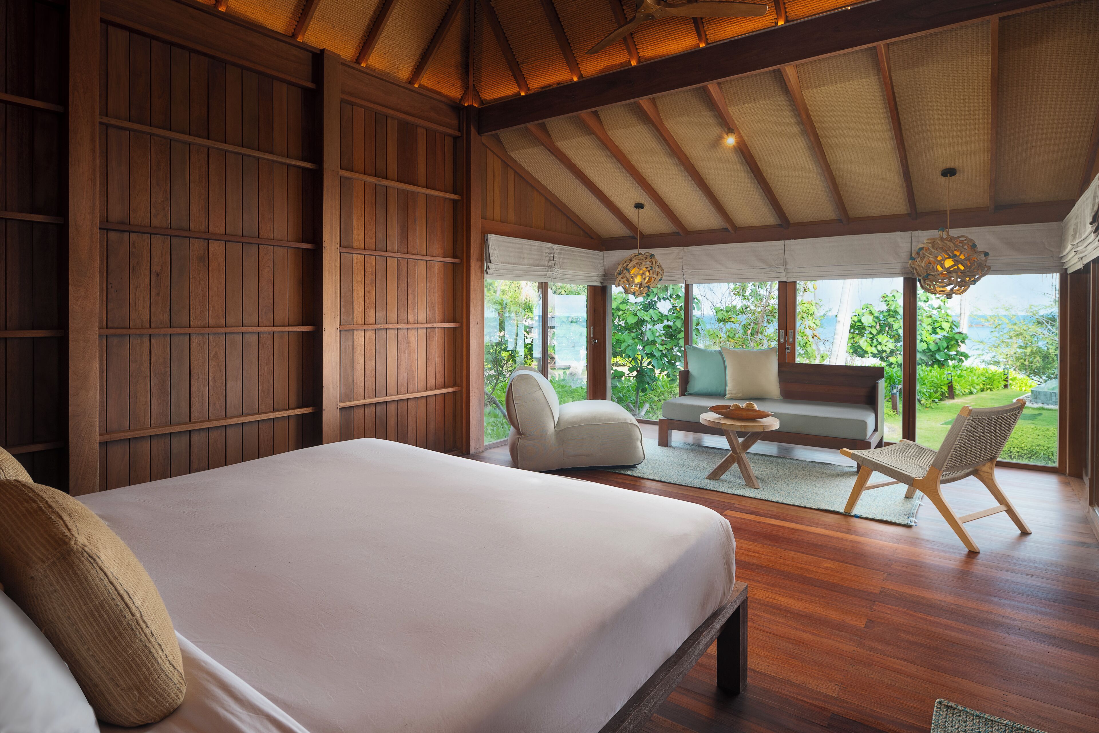 Un dormitorio con una cama grande, una estantería de madera, una zona de estar con sofá y sillones, y vistas a la vegetación a través de grandes ventanales.