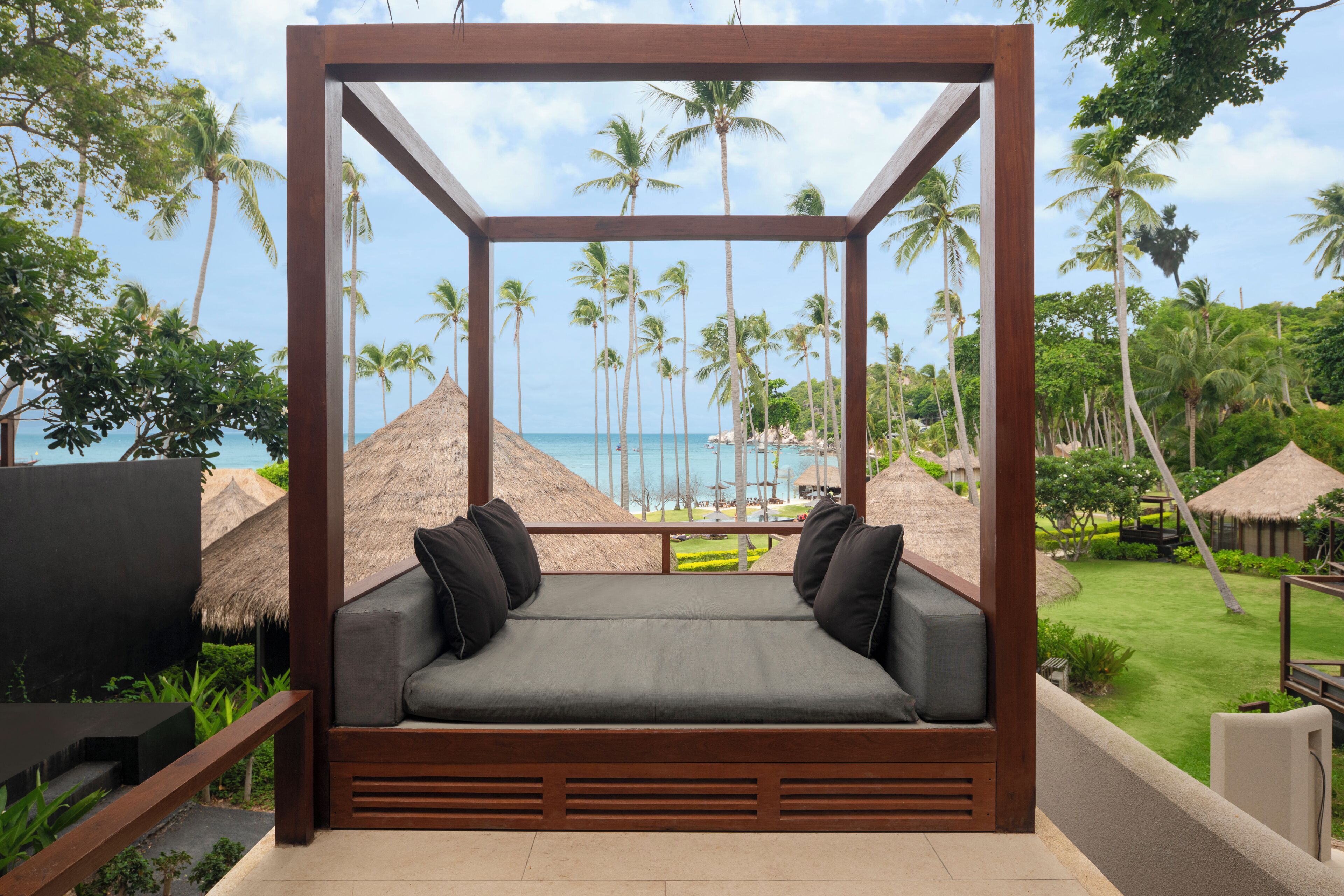 A wooden-framed outdoor seating area with a cushioned bench and pillows, overlooking a tropical beach with palm trees and a clear sky.