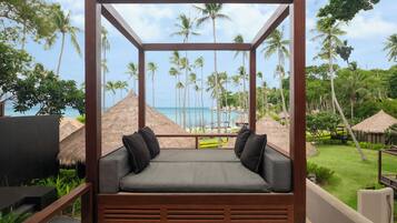 A wooden-framed outdoor seating area with a cushioned bench and pillows, overlooking a tropical beach with palm trees and a clear sky.