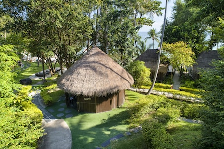 A thatched-roof hut with a paved path leading to it, surrounded by lush greenery and palm trees.