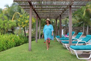 Una mujer caminando bajo una pérgola de madera en un jardín con césped verde, palmeras y un área de piscina con sillones de descanso.
