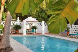 Piscina con sillones de descanso y una mesa, rodeada por una valla blanca y follaje.