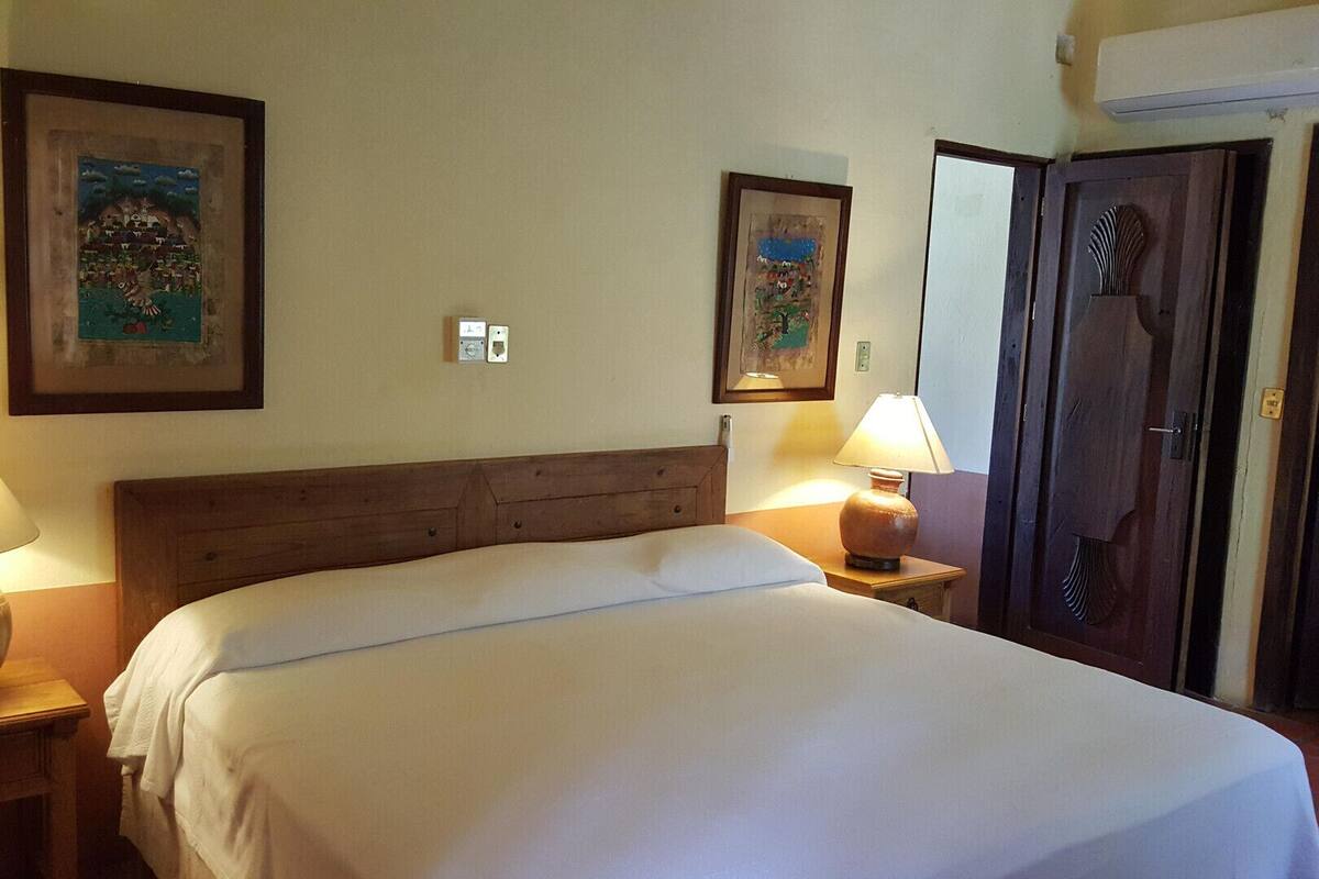 A hotel room with a bed, two bedside lamps, a wall-mounted air conditioner, and two framed pictures.