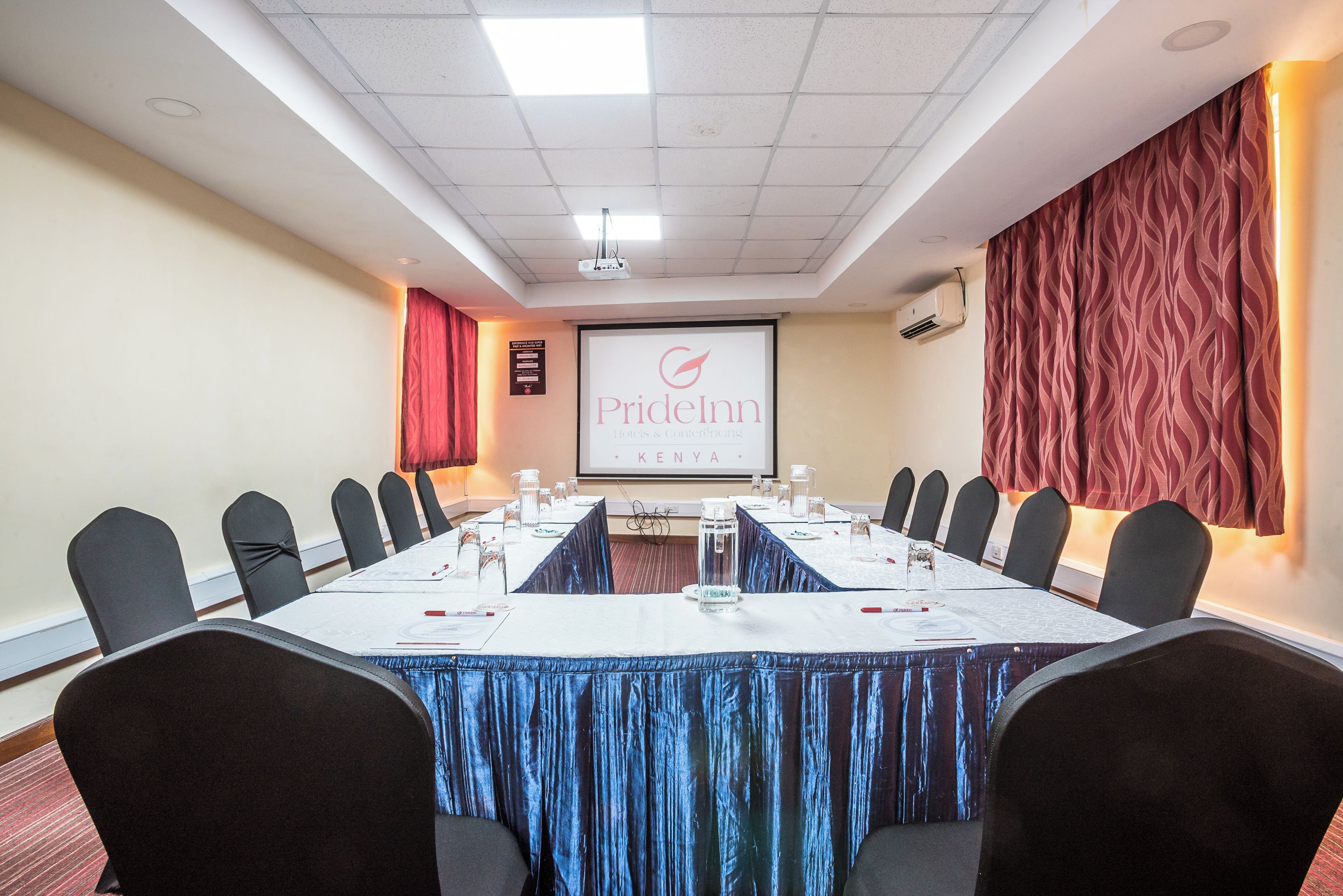 Un salón de conferencias con una larga mesa, sillas, una pantalla de proyector y una cortina roja.
