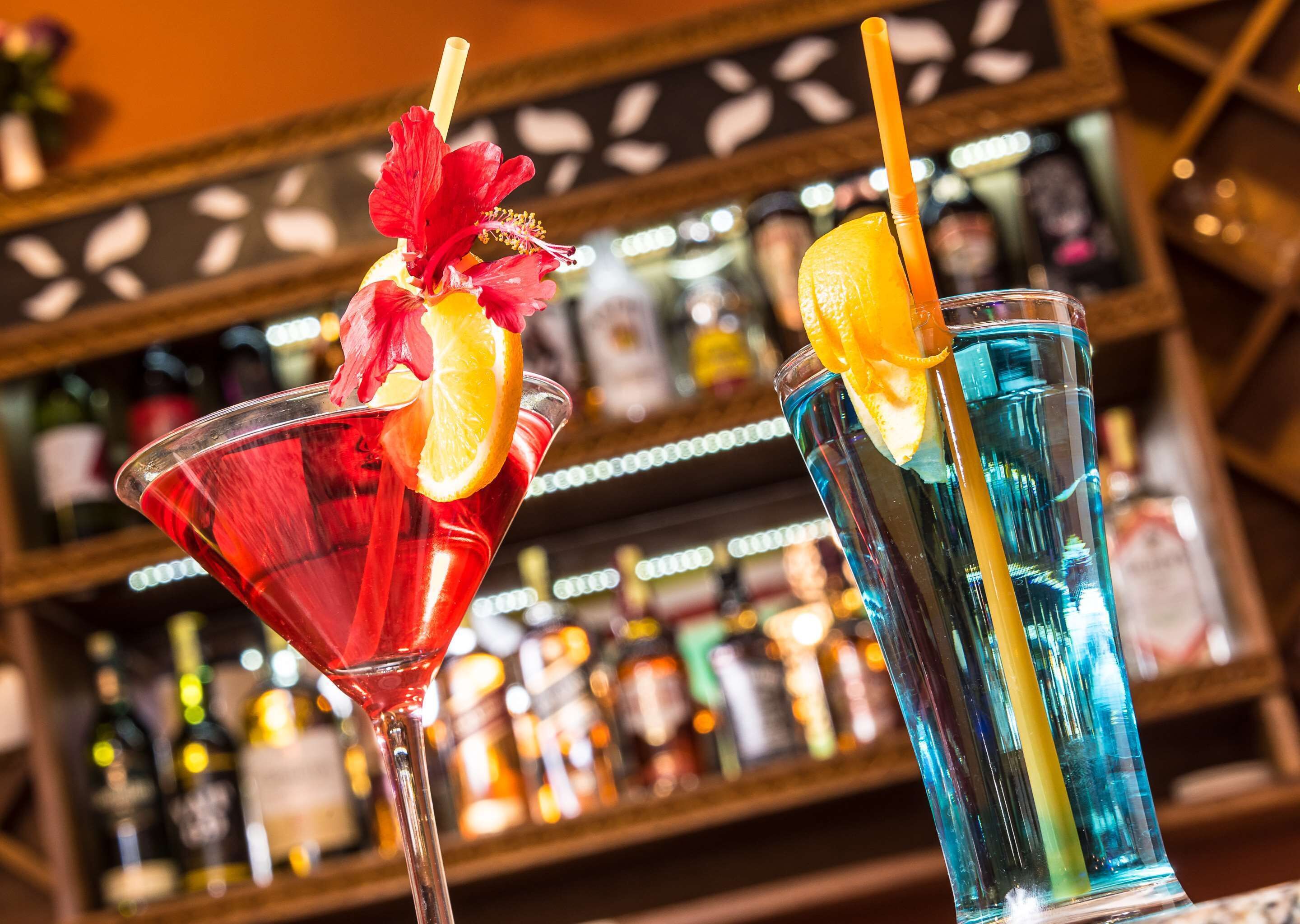 Two cocktails with garnishes and straws, placed on a bar counter with bottles in the background.