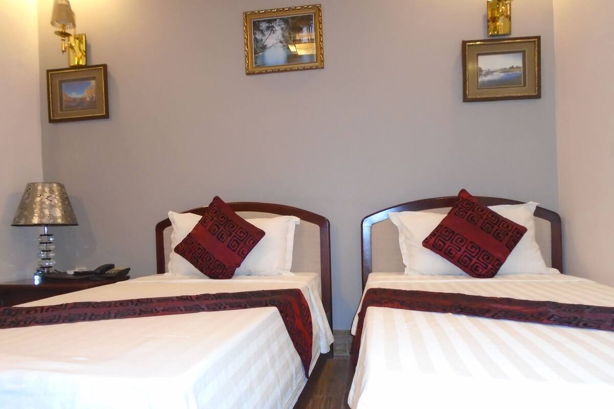 Two single beds with white linens and red patterned pillows, a nightstand with a lamp, and framed pictures on the wall.