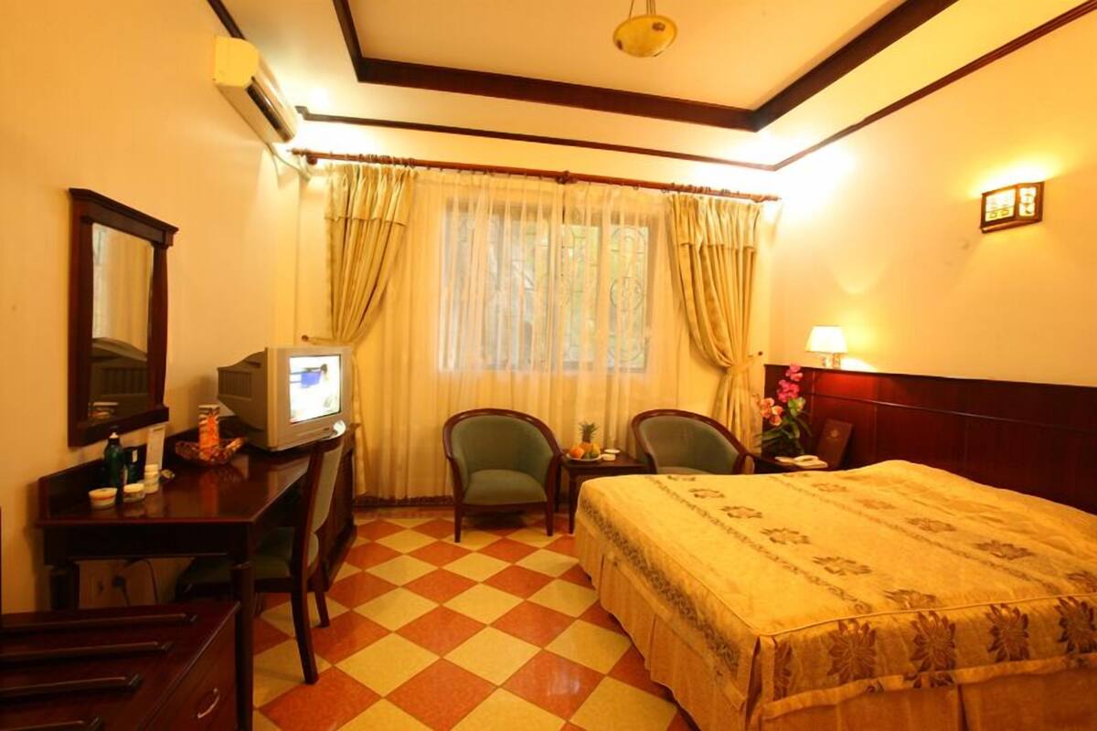 A hotel room with a bed, two chairs, a desk with a computer, a small table with a lamp, and a window with curtains.
