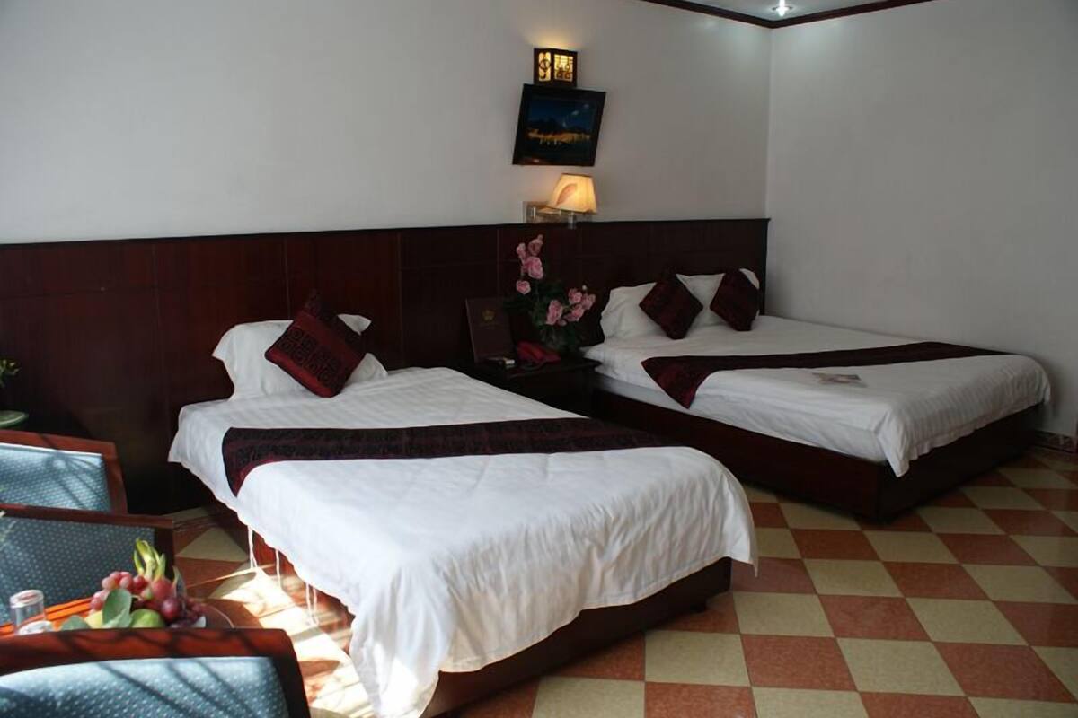 A hotel room with two single beds, a nightstand, a lamp, a framed picture, and a small table with fruit.