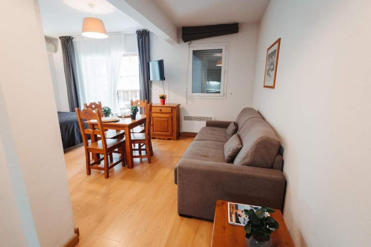 A cozy living room with a brown sofa, wooden dining table, and chairs. There is a TV mounted on the wall and a window with curtains.