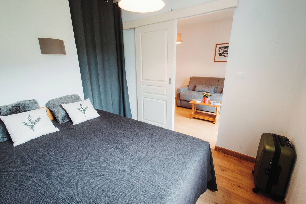 A bedroom with a large bed, a suitcase, and a view into a living area with a sofa and a table.