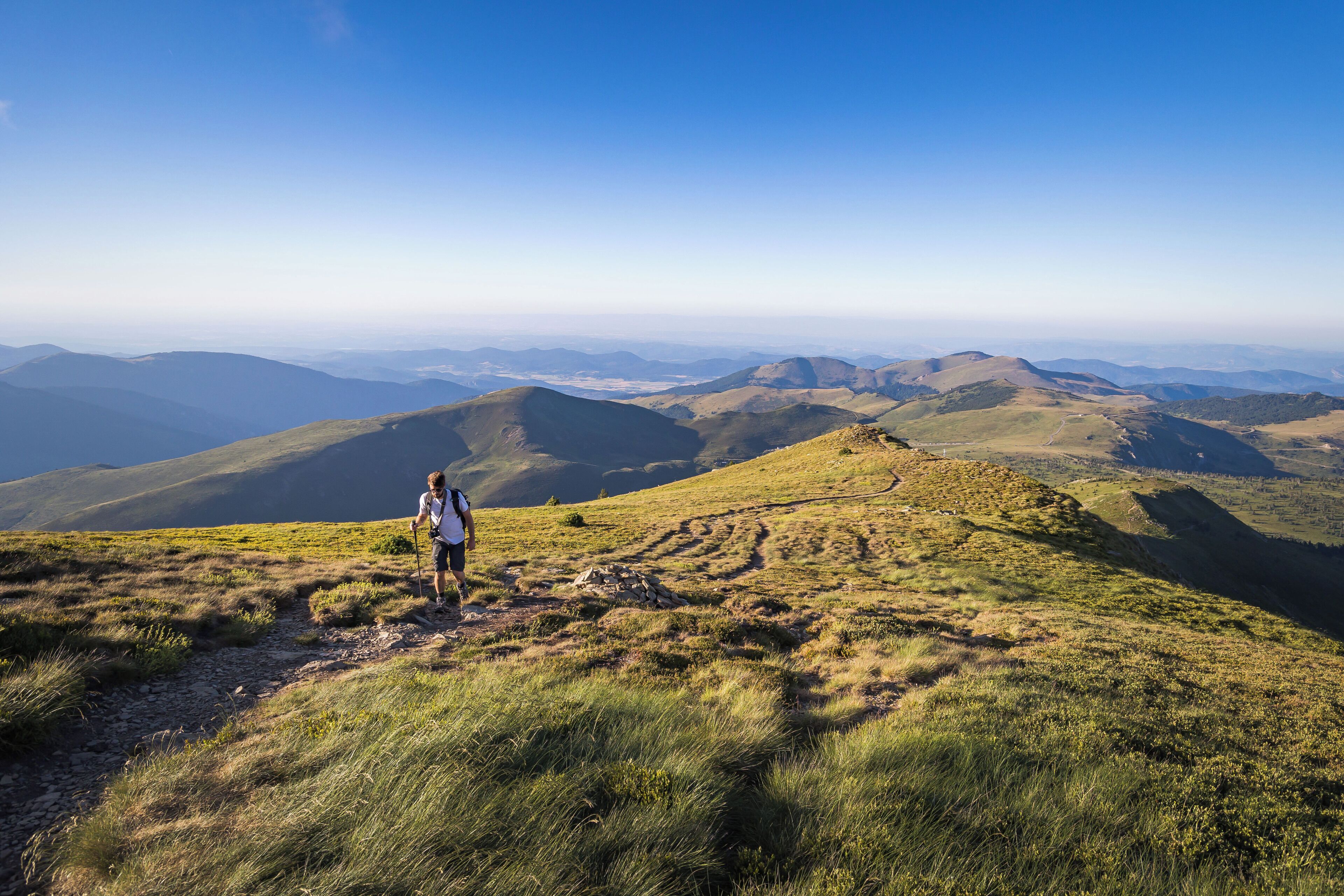Un escursionista su un sentiero montano erboso con viste panoramiche su colline e valli lontane.