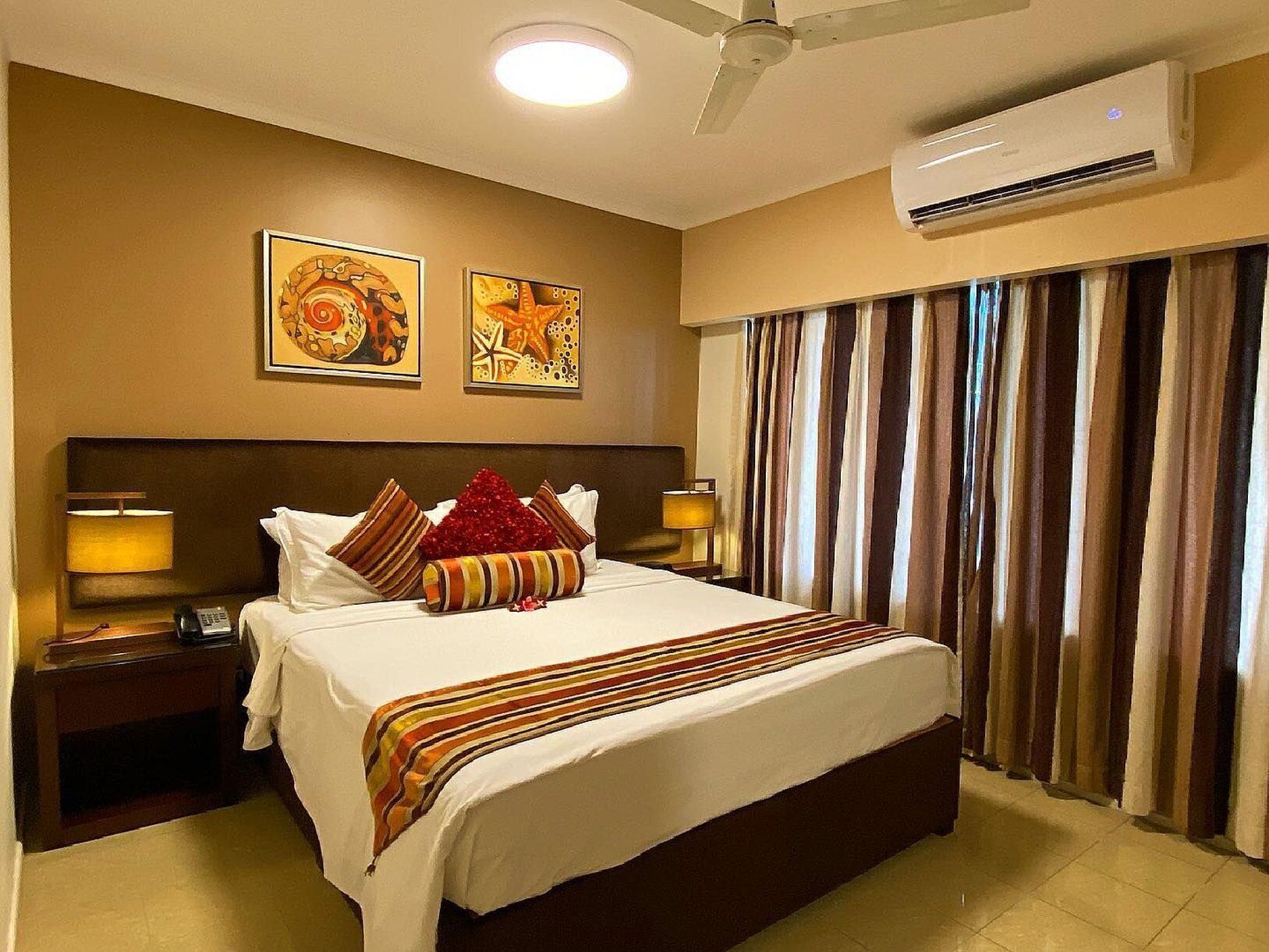 A hotel room with a large bed, two wall-mounted paintings, a ceiling fan, an air conditioning unit, and a window with striped curtains.