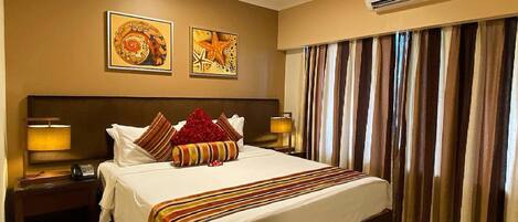 A hotel room with a large bed, two wall-mounted paintings, a ceiling fan, an air conditioning unit, and a window with striped curtains.