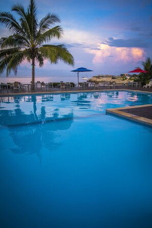 A swimming pool with palm trees and beach chairs, surrounded by umbrellas. - Heritage Park Hotel (Honiara)