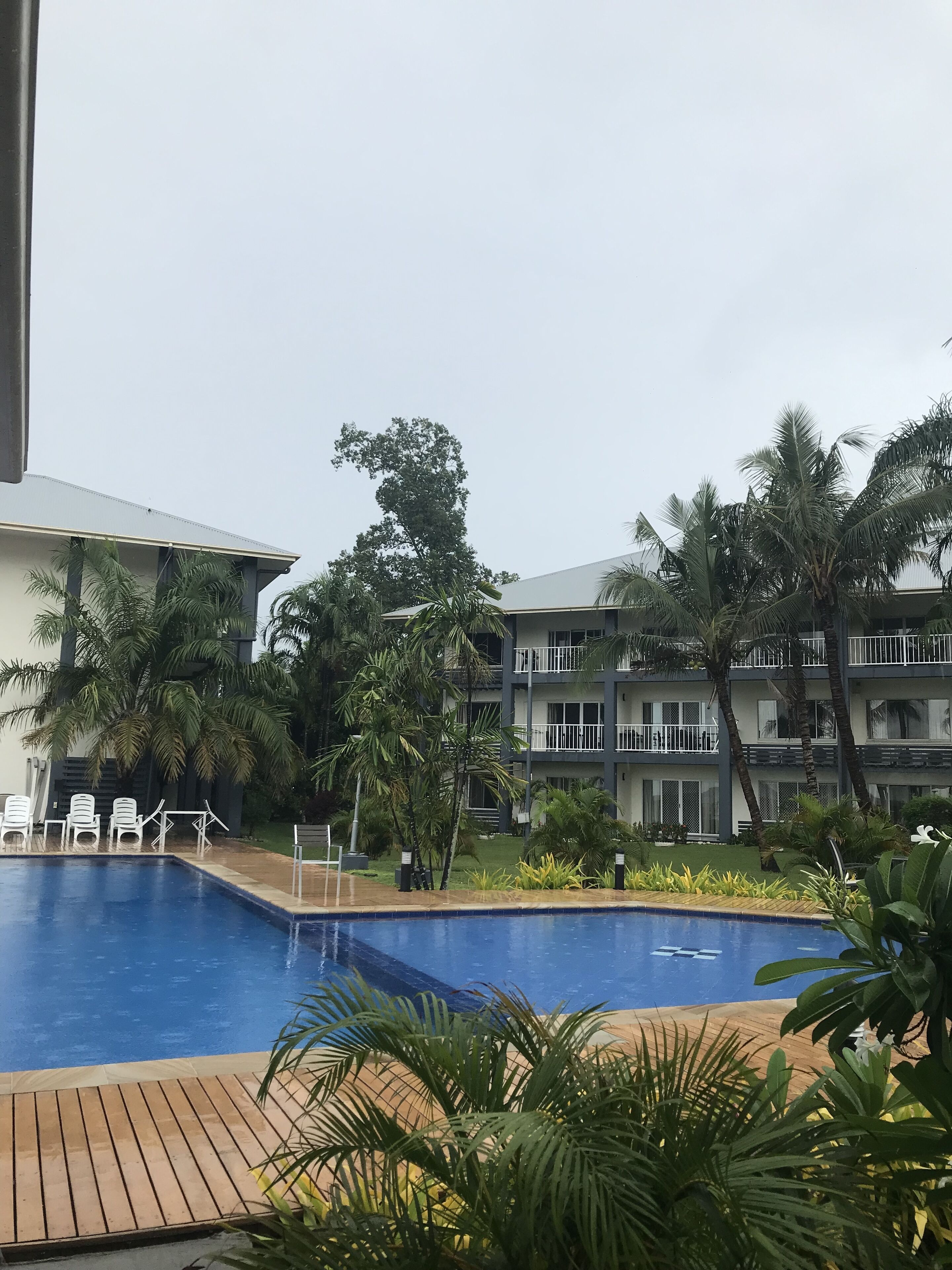 A swimming pool with a wooden deck, surrounded by palm trees and a multi-story building with balconies.