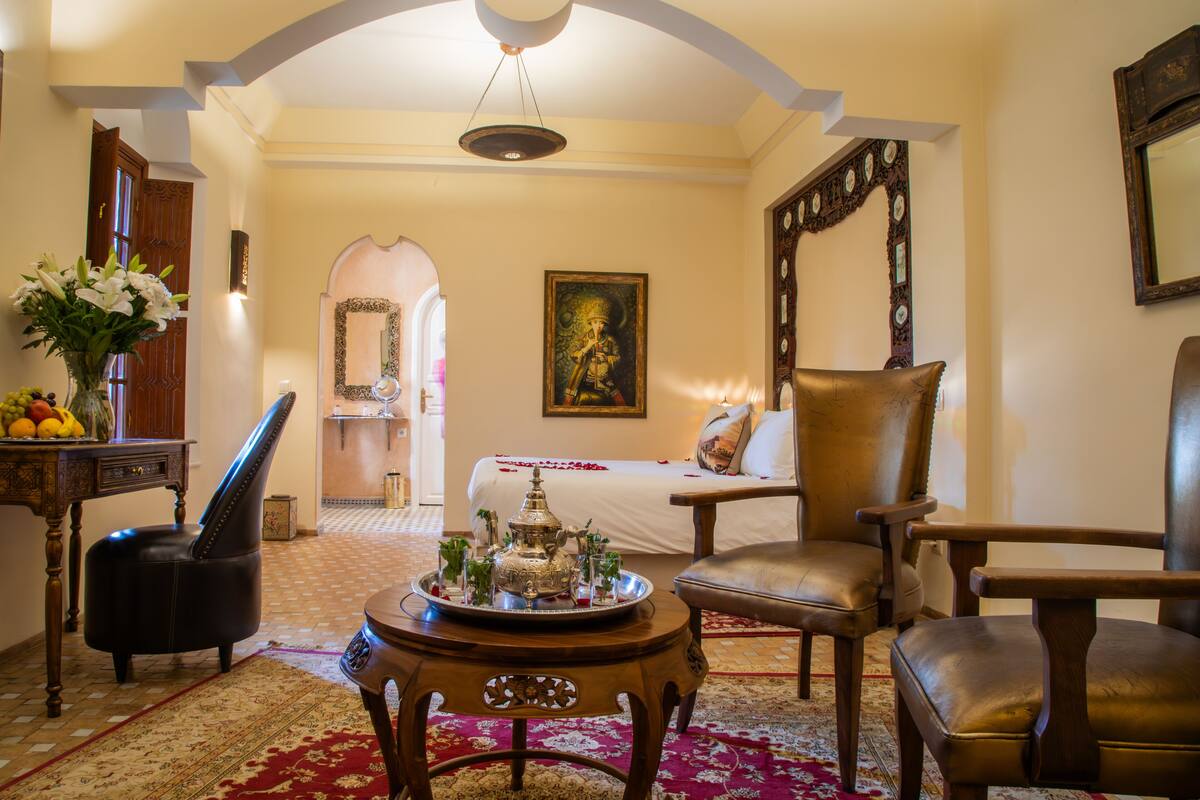 A traditional room with a bed, two leather chairs, a wooden side table with a tray of items, a mirror, and a painting on the wall.