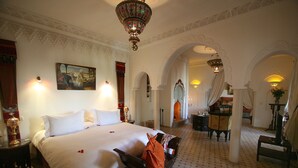 A hotel room with a large bed, a bench, a desk, and a chair. There are two lamps on the wall and a chandelier. - Demeures d'orient Riad Deluxe & Spa (Marrakech)