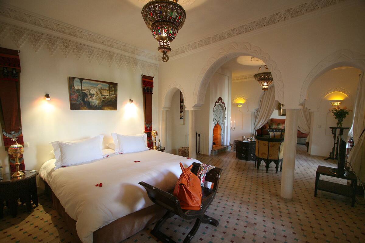 A hotel room with a large bed, a bench, a desk, and a chair. There are two lamps on the wall and a chandelier.