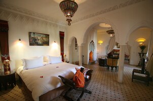 Habitación de hotel con una cama grande, una banca, un escritorio y una silla. Hay dos lámparas en la pared y un candelabro.