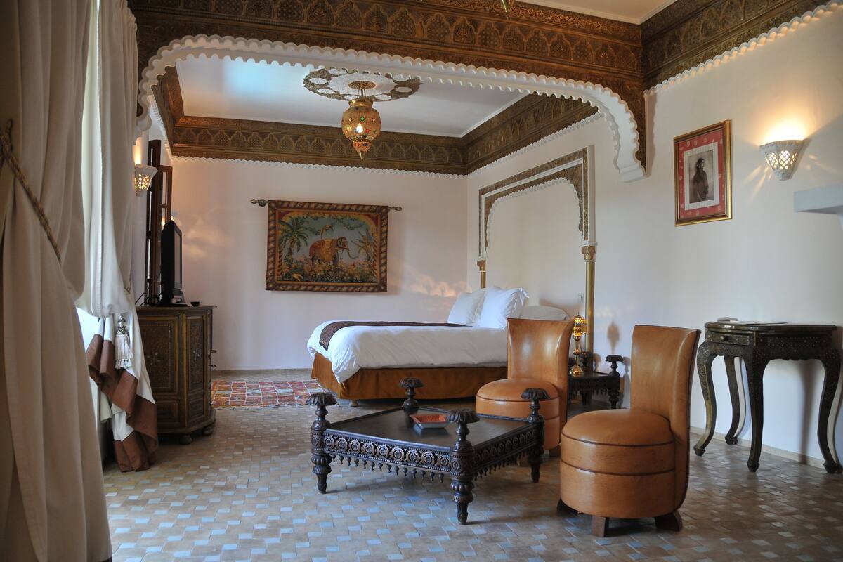 A traditional room with a four-poster bed, a wooden chair, a small table, and a decorative ceiling.