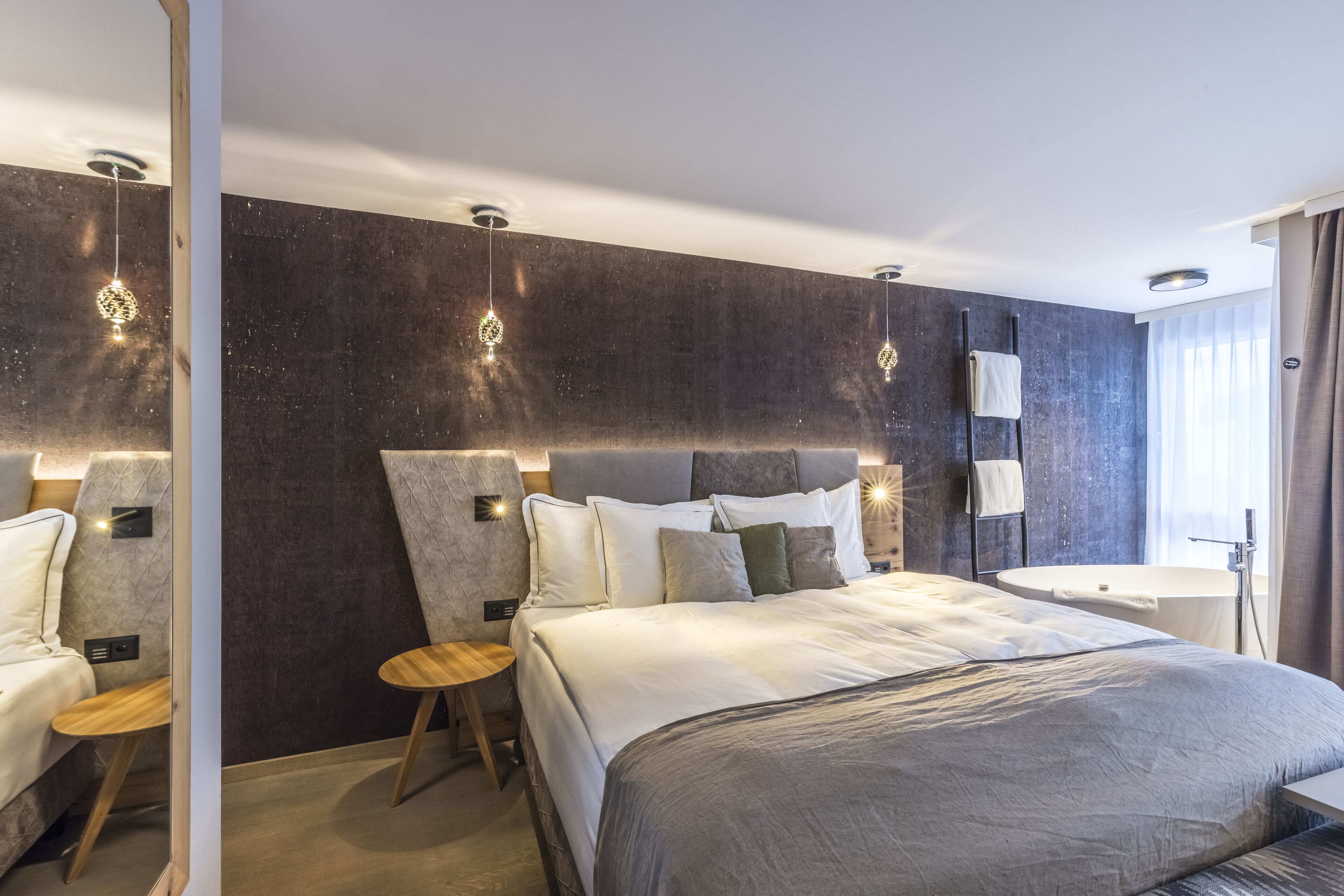 A modern hotel room with a large bed, bedside tables, a vanity with a mirror, and a bathtub.