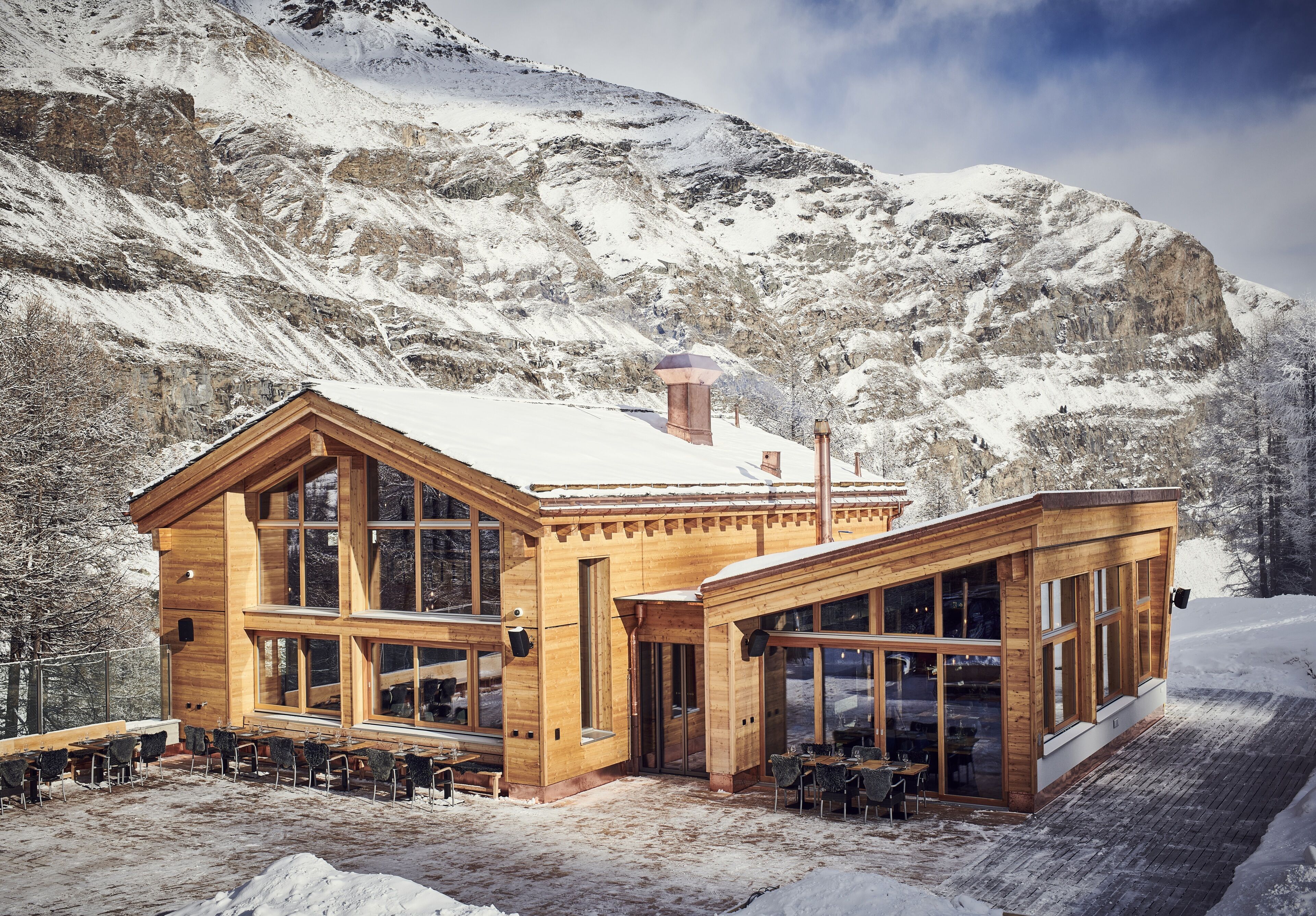 A wooden chalet with large windows and a snow-covered roof, situated in a mountainous area.